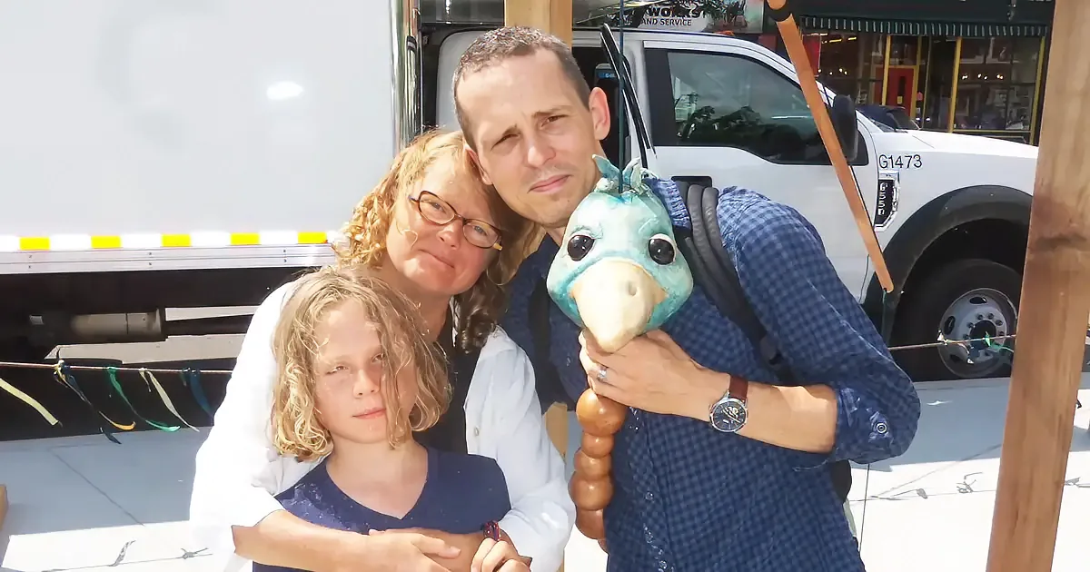 A family of three poses together, the father holding a blue stuffed animal, with a white truck and colorful ribbons in...