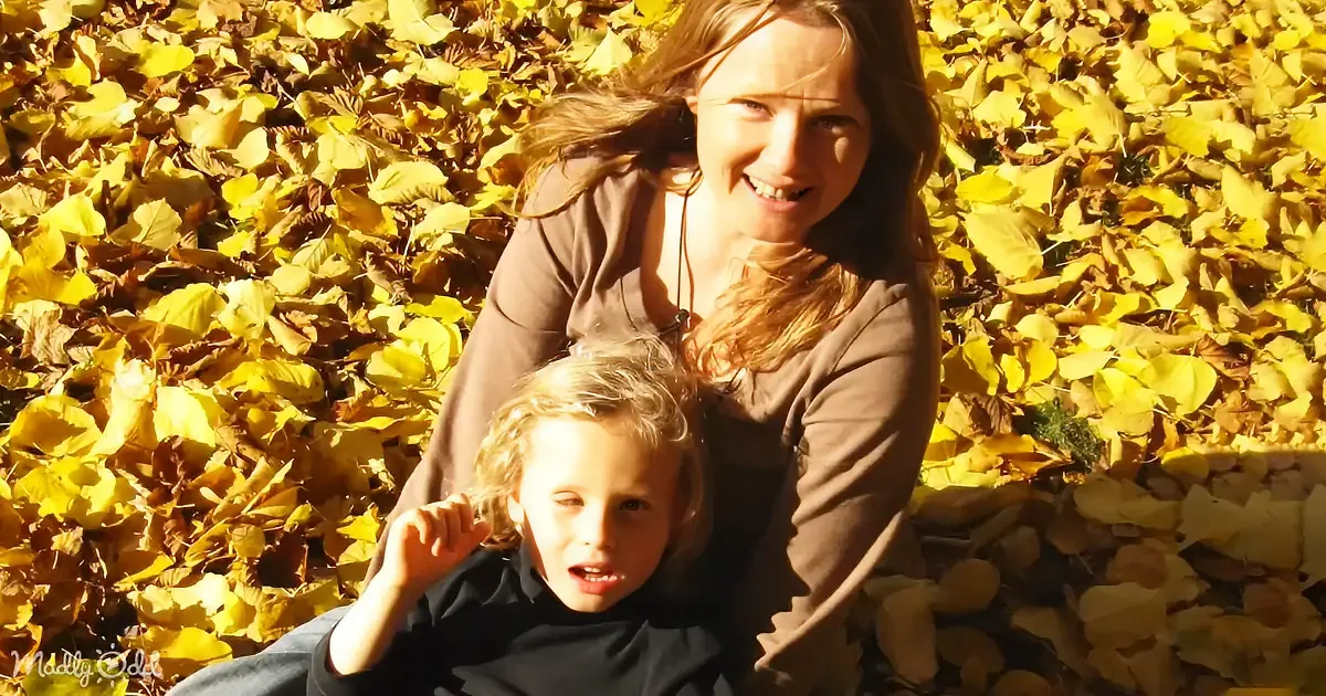 A woman and child sit amidst golden autumn leaves, smiling and enjoying the sunny day.