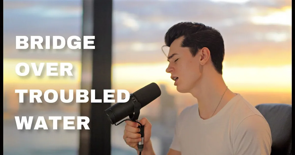 A person sings into a microphone, wearing a white shirt, with a sunset view through the window behind them.