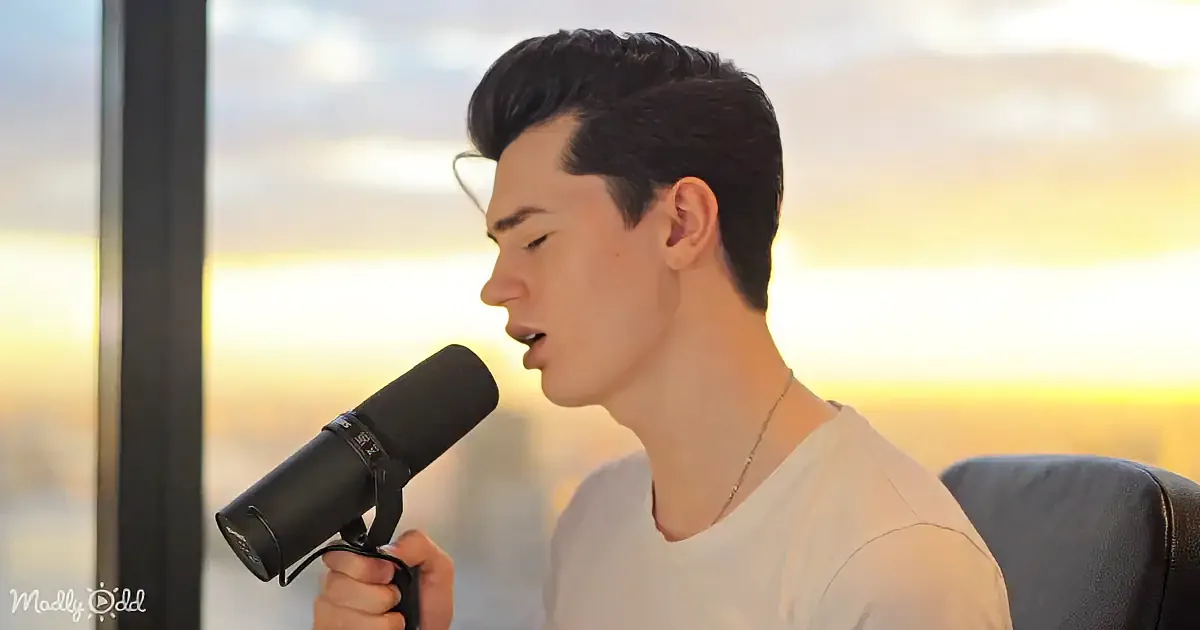A person sings into a microphone, wearing a white shirt, with a sunset view in the background.