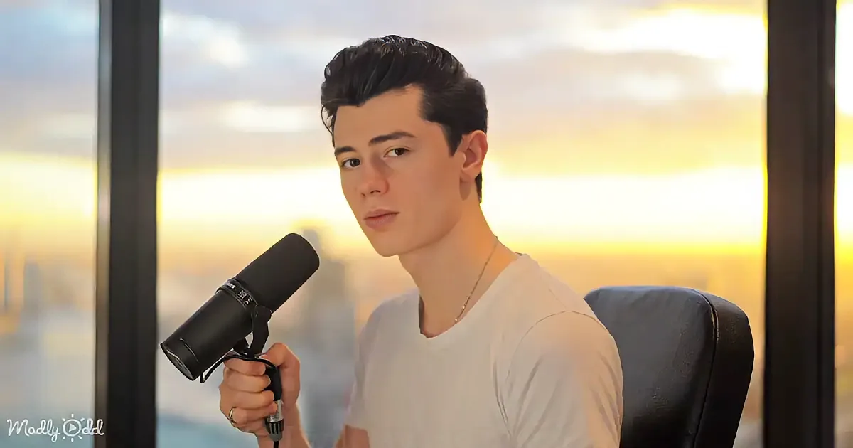 A person in a white shirt holds a microphone, sitting in front of a window with a cityscape and sunset view.