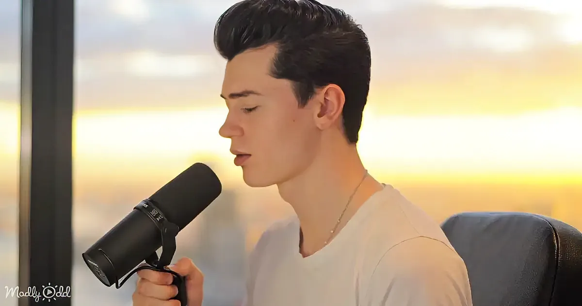A person in a white shirt holds a microphone, seated against a sunset backdrop.