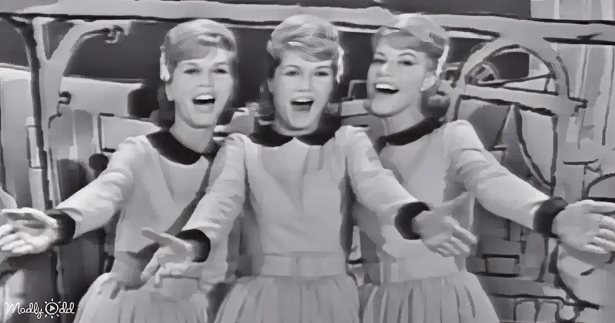 Three women in matching dresses sing and gesture joyfully, arms outstretched, in a black-and-white scene.