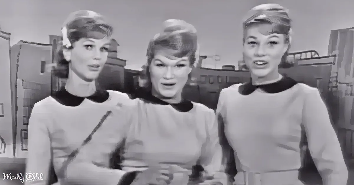Three women in matching outfits stand together, possibly singing or speaking, in a black-and-white setting.