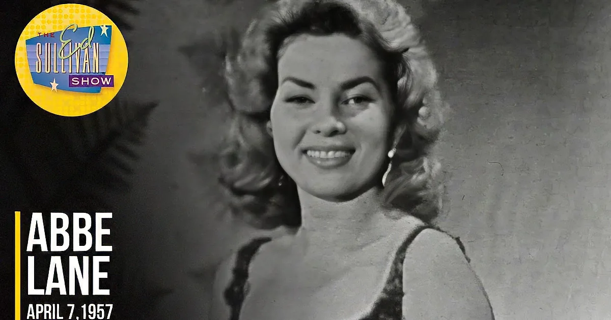 A smiling woman with curly hair, wearing a sleeveless top, stands in front of a decorative plant.