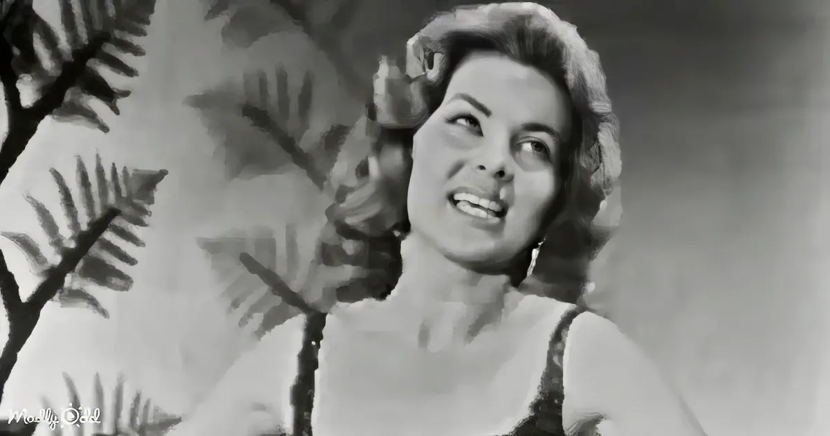 A woman with curly hair and earrings, wearing a sleeveless top, looks upwards with a tropical plant in the background.