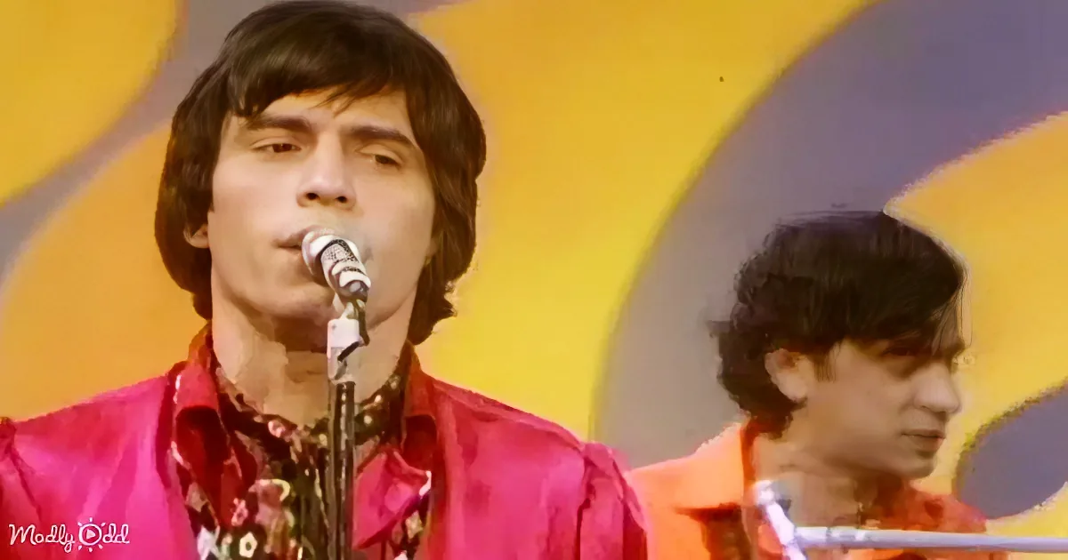 A man in a red jacket sings into a microphone, with another person in the background, both against a colorful backdrop.