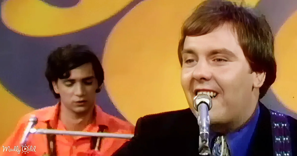 A man in a suit sings into a microphone, with another person in an orange shirt behind him.