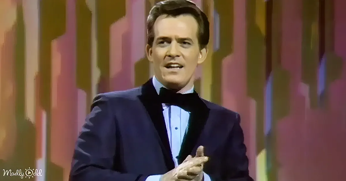 A man in a dark suit and bow tie gestures while speaking on stage.