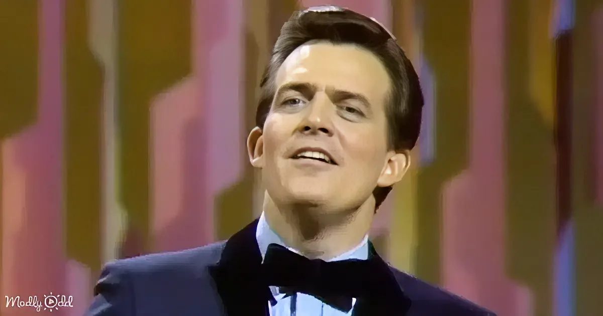 A man in a tuxedo with a bow tie, smiling, against a colorful striped background.