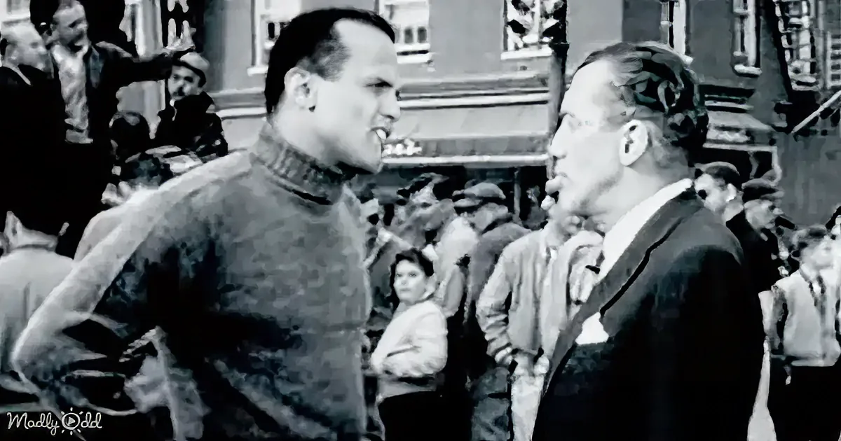 Two men face each other in a crowded street scene, one in a sweater, the other in a suit, surrounded by onlookers.