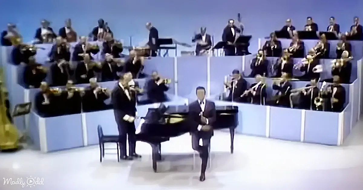 A man in a suit plays piano in front of a large orchestra in a studio setting.