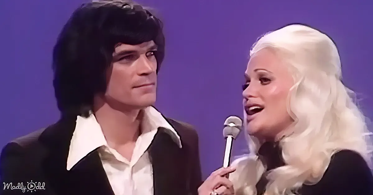 A man in a black suit and a woman with long blonde hair holding a microphone are on stage.