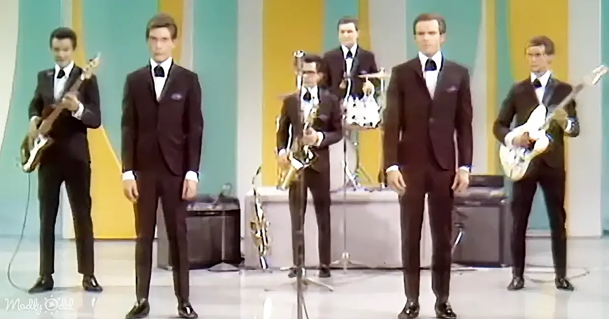 A band in suits performs on stage with a saxophone, guitars, and a bass.