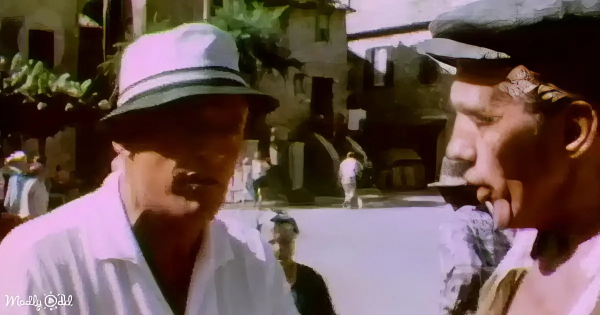 Two men in hats and white shirts are conversing, with a blurred background of a building and other people.