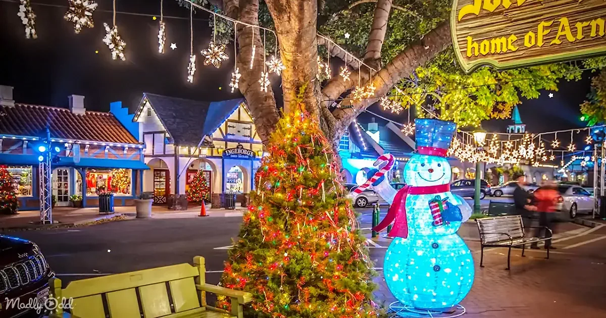 A brightly lit snowman statue stands beside a decorated Christmas tree in a festive town square at night.