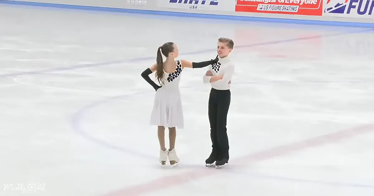 A figure skating pair in white and black costumes stands on ice, holding hands, with a US Figure Skating banner in the...