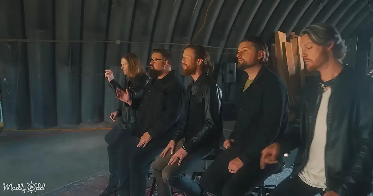 Five men in black leather jackets sit in a row, facing left, in a dimly lit, industrial setting.