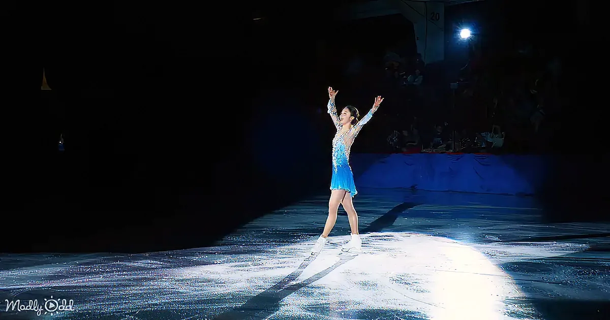 A figure skater in a blue dress stands on ice, arms raised, spotlight illuminates her.