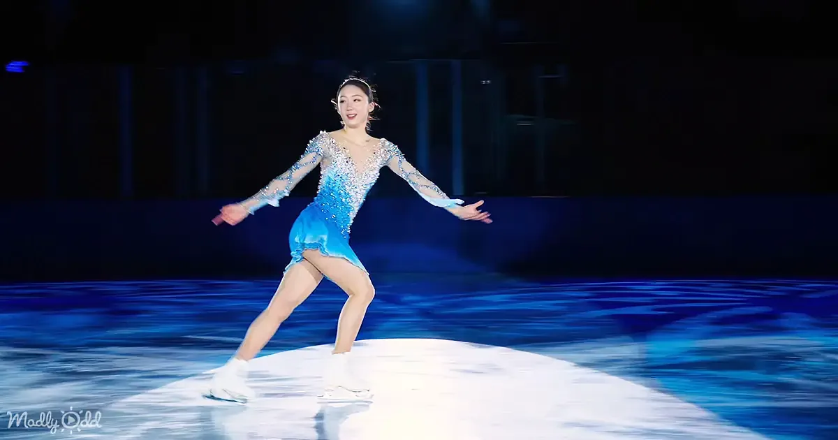 A figure skater in a blue dress glides gracefully on ice, arms outstretched.