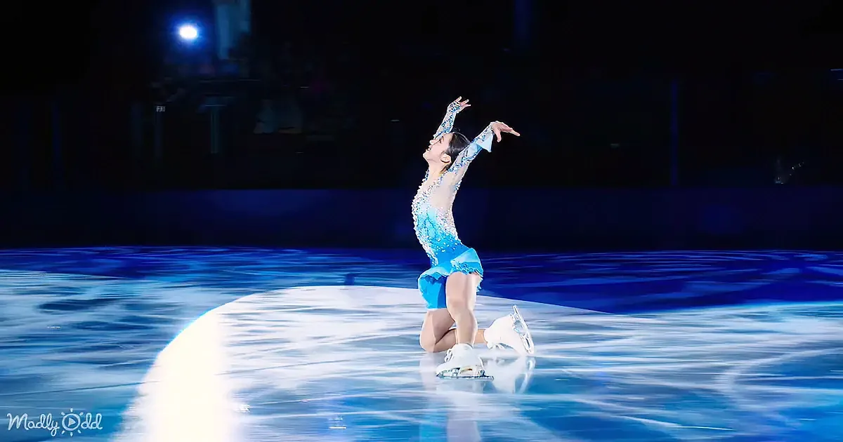 A figure skater in a blue outfit kneels on one knee on the ice, arms raised, illuminated by stage lights.