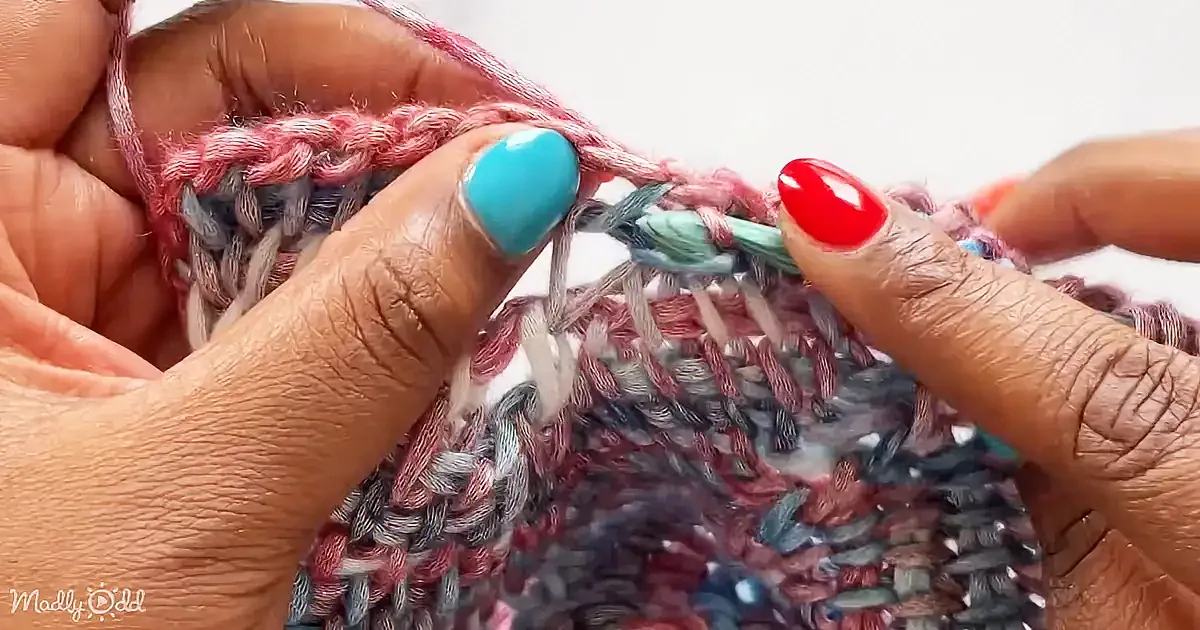 Hands with colorful nails hold a partially completed knitted fabric with pink and gray yarn.