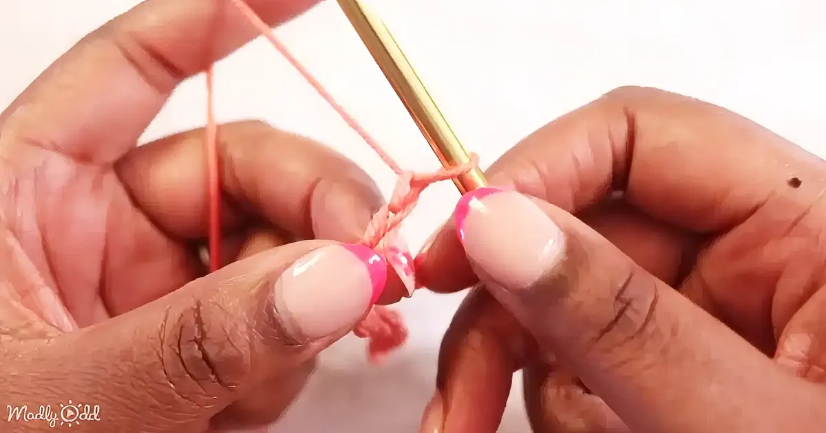 Hands with pink nails hold a crochet hook and pink yarn, demonstrating a crochet technique.