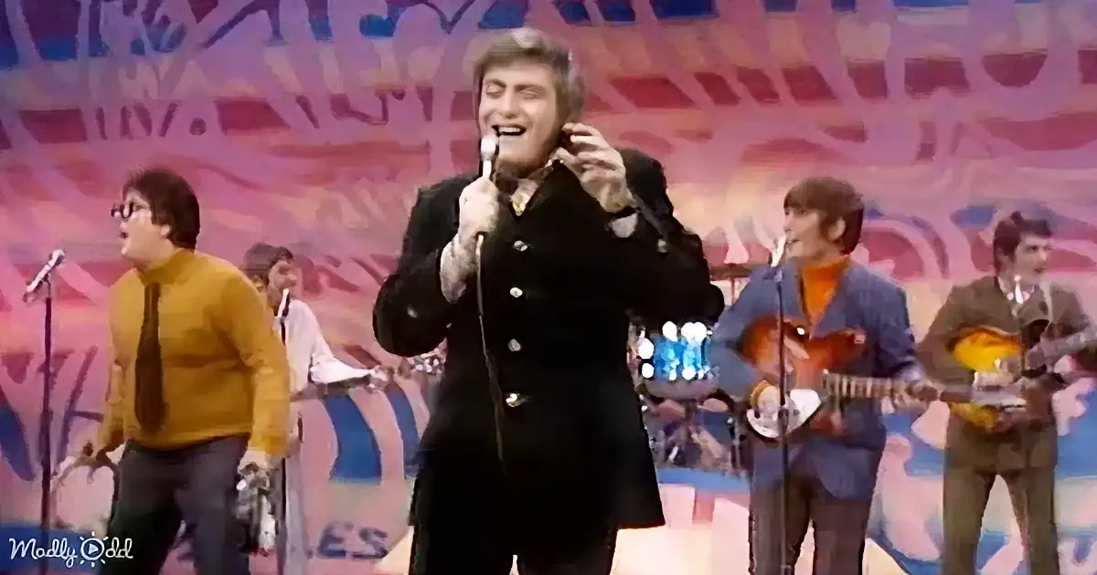 A man in a black coat sings into a microphone on stage with a colorful backdrop and band.