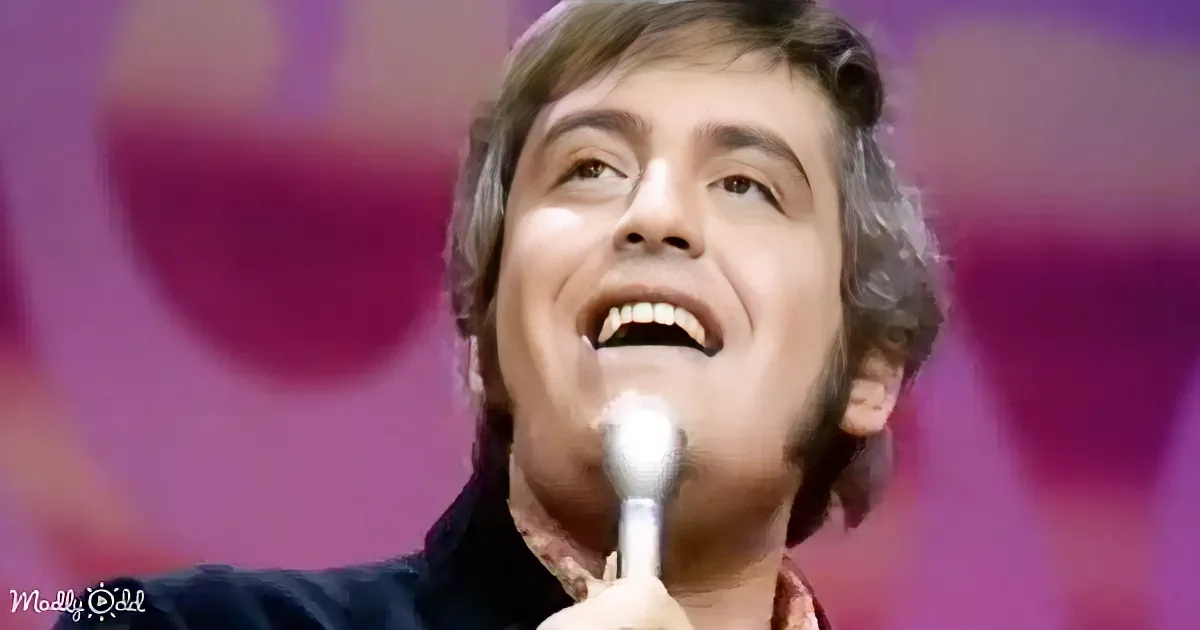 A man with curly hair singing into a microphone, wearing a dark jacket and a red shirt, against a blurred pink background.