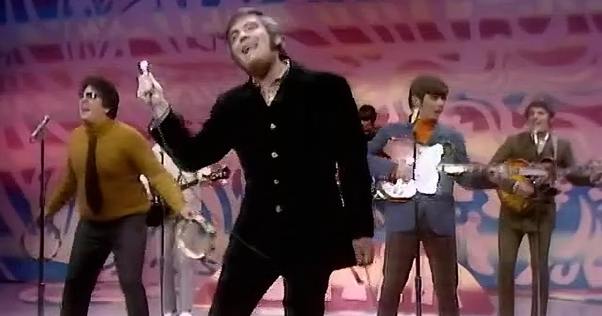 A man with dark hair and a smile holds a microphone, wearing a dark outfit and a necklace, against a blurred purple...