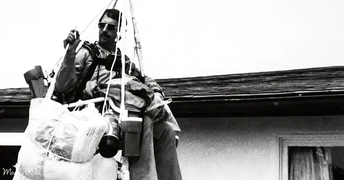 A man in a harness and helmet is suspended in mid-air, holding onto ropes, with a building and sky in the background.