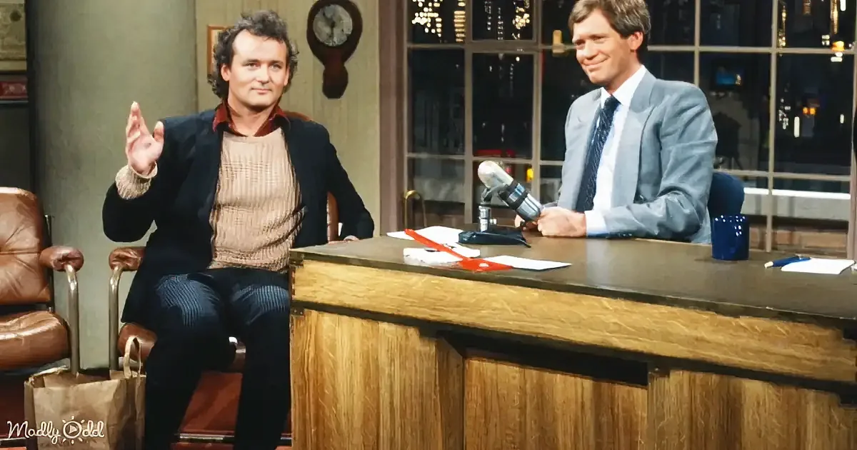 A man in a sweater and blazer gestures while seated, with another man in a suit behind him at a desk.