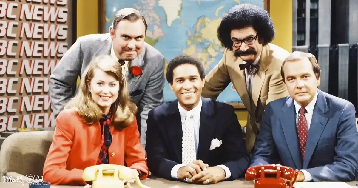 A news anchor and his team, including a woman in a red blazer, are seated at a desk with a world map in the background.