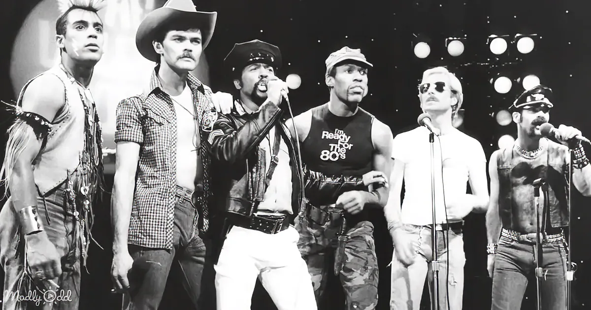 A man in a leather jacket and hat sings into a microphone, surrounded by other performers in various costumes.