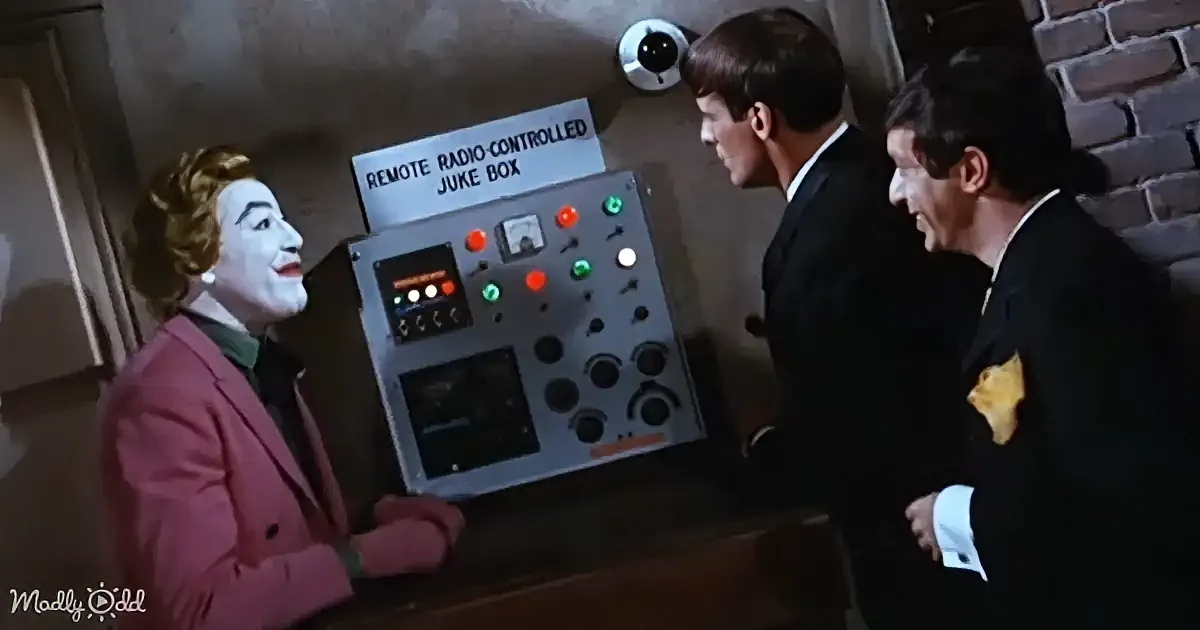 The Joker in a pink suit stands beside a remote radio-controlled jukebox, interacting with two men in suits.