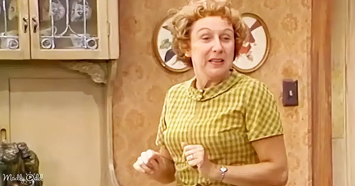 A woman in a yellow checkered shirt stands in a kitchen, holding a white cloth, with patterned wallpaper and cabinets in...