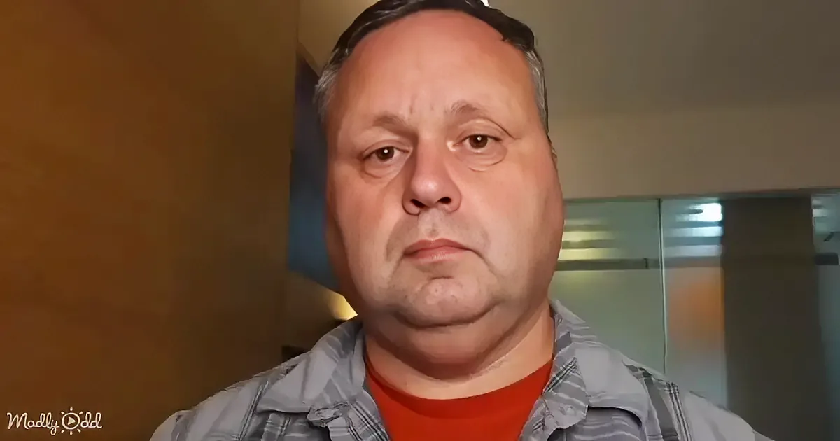 A man with short dark hair, wearing a plaid shirt over a red t-shirt, looks directly at the camera.