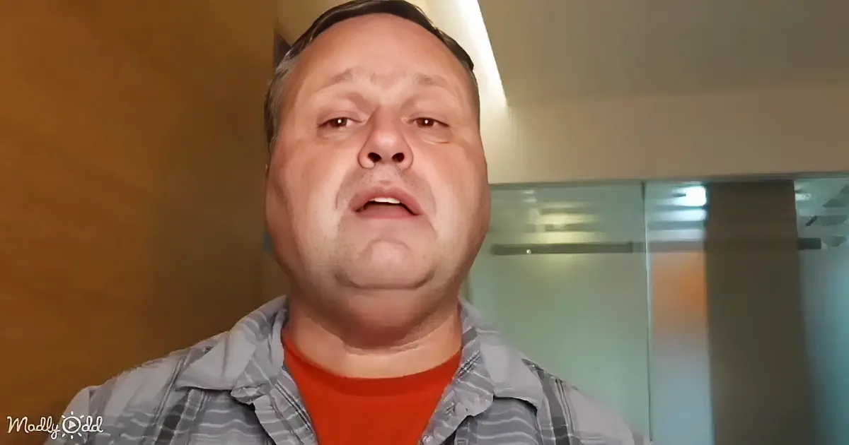 A man wearing a striped shirt and red undershirt looks upwards with a neutral expression.