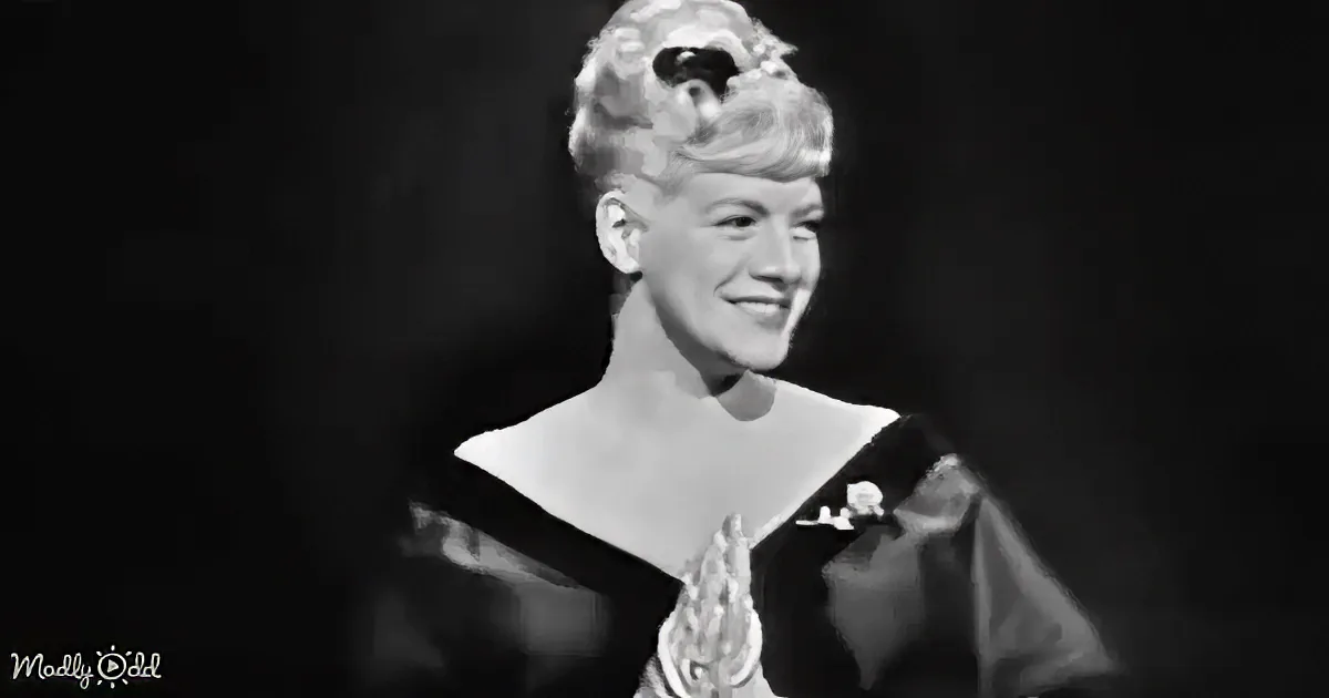 A woman with blonde hair styled in curls, wearing a black dress, stands against a blurred background.