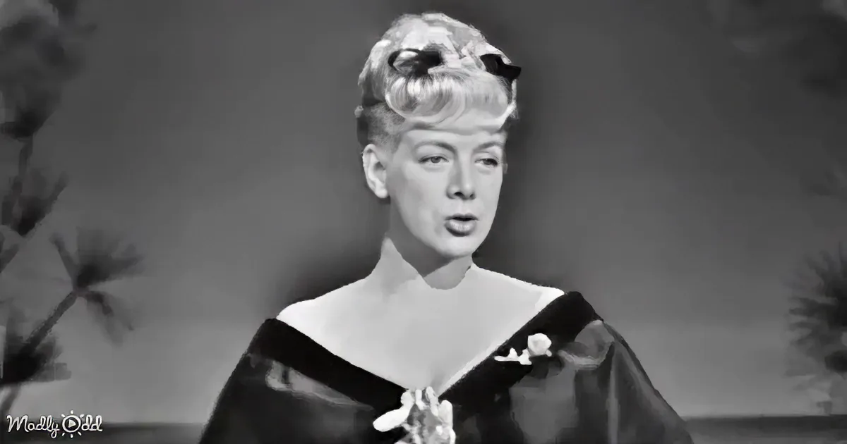 A woman in a black dress with a floral headpiece and a flower on her chest, standing against a plain background.