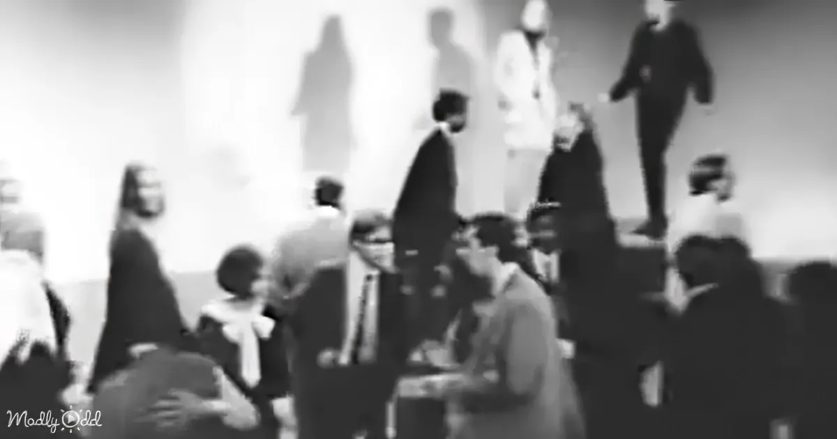A blurry black-and-white image shows a group of people, some standing and some sitting, in what appears to be a public...