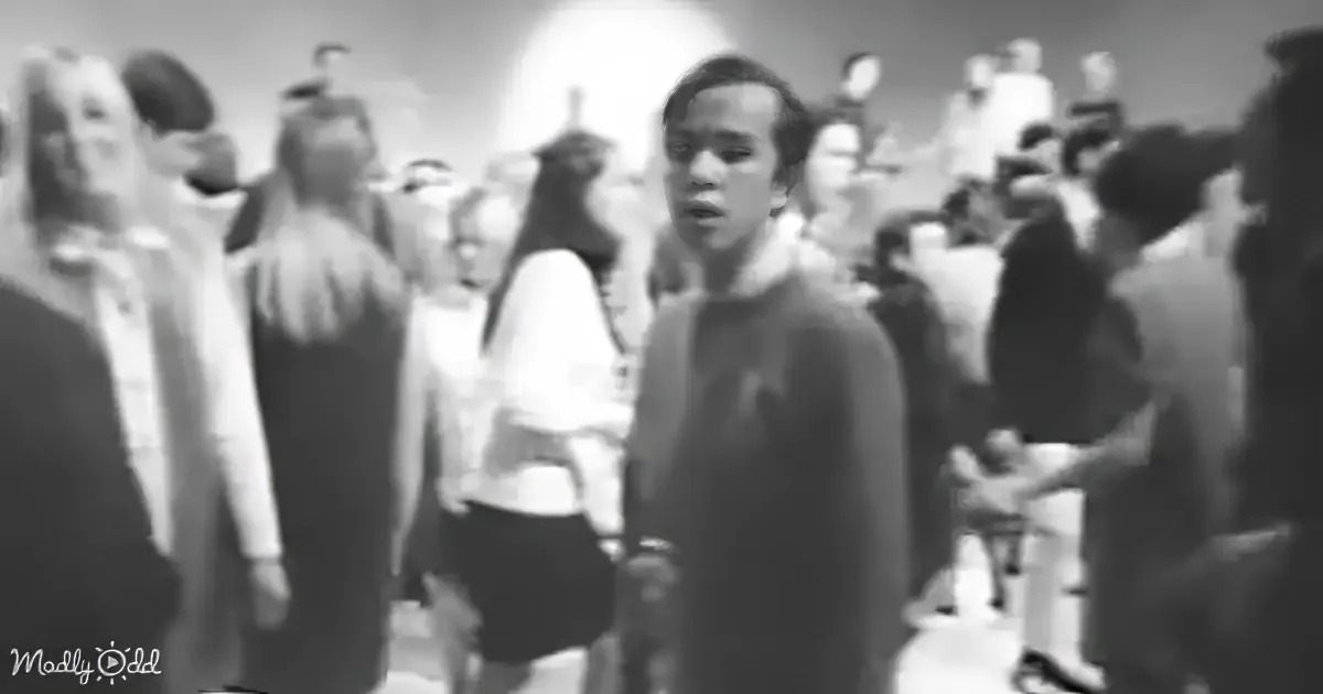 A man in a dark coat stands in a crowded, blurry scene, surrounded by people in various attire.