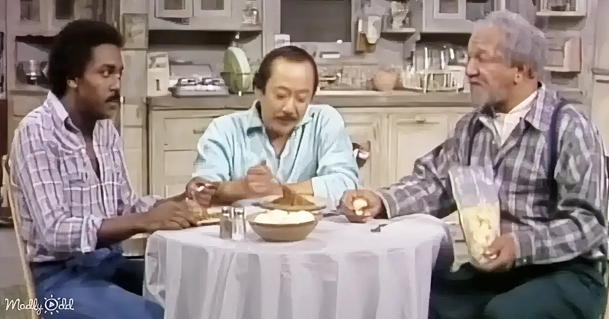 Three men sit at a kitchen table, eating and conversing, with one holding popcorn.