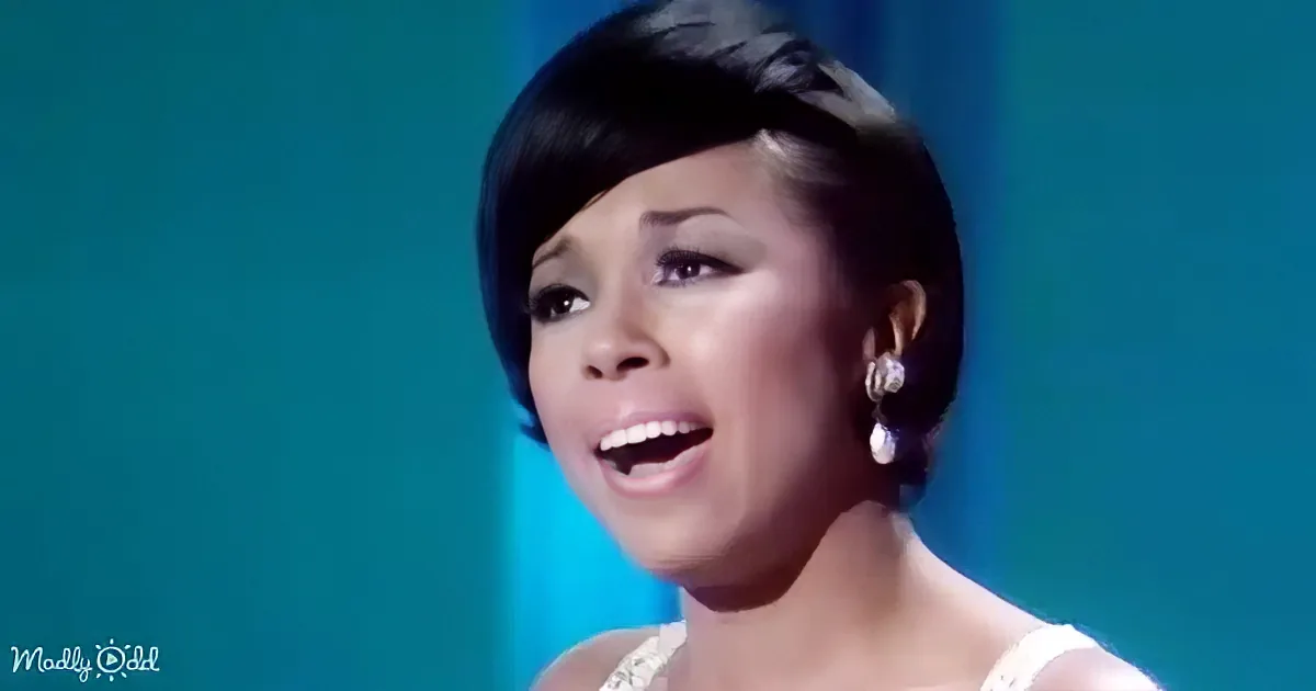 A woman with short black hair and earrings is singing, wearing a white outfit against a teal background.