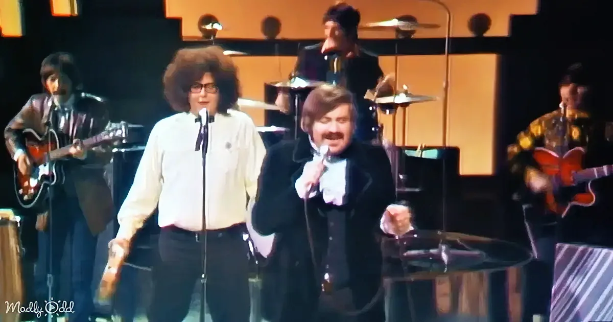A man in a black coat and white shirt sings into a microphone on stage with a band.