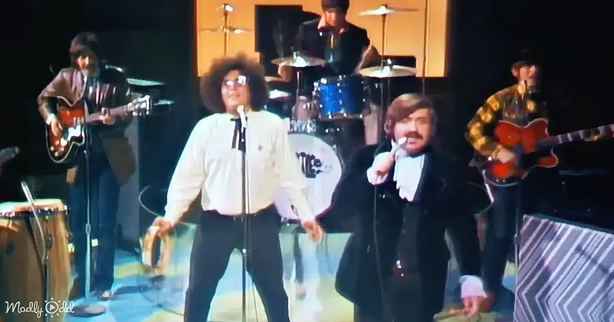 Two men sing on stage with a band, one in a white shirt, the other in a black suit.