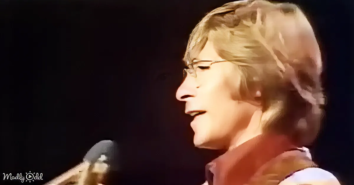 A person with blonde hair and glasses is singing into a microphone, illuminated by stage lighting.