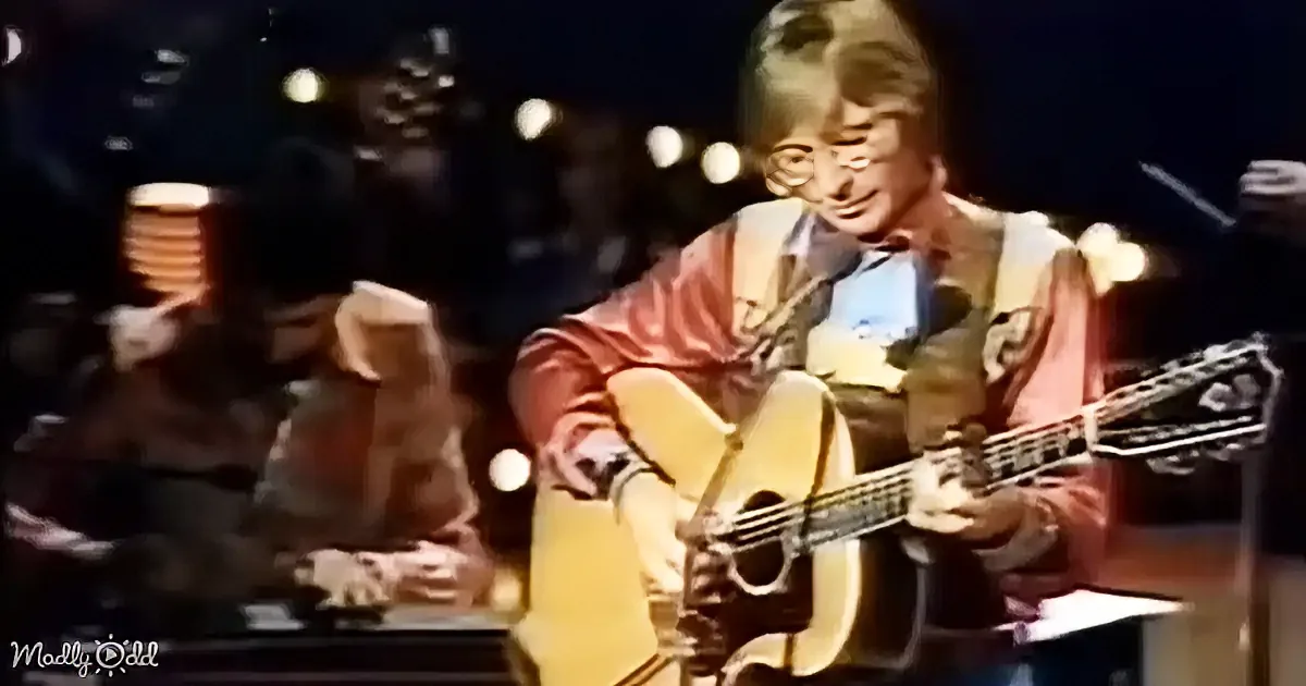 A person in a red jacket plays an acoustic guitar on stage with a microphone in front.