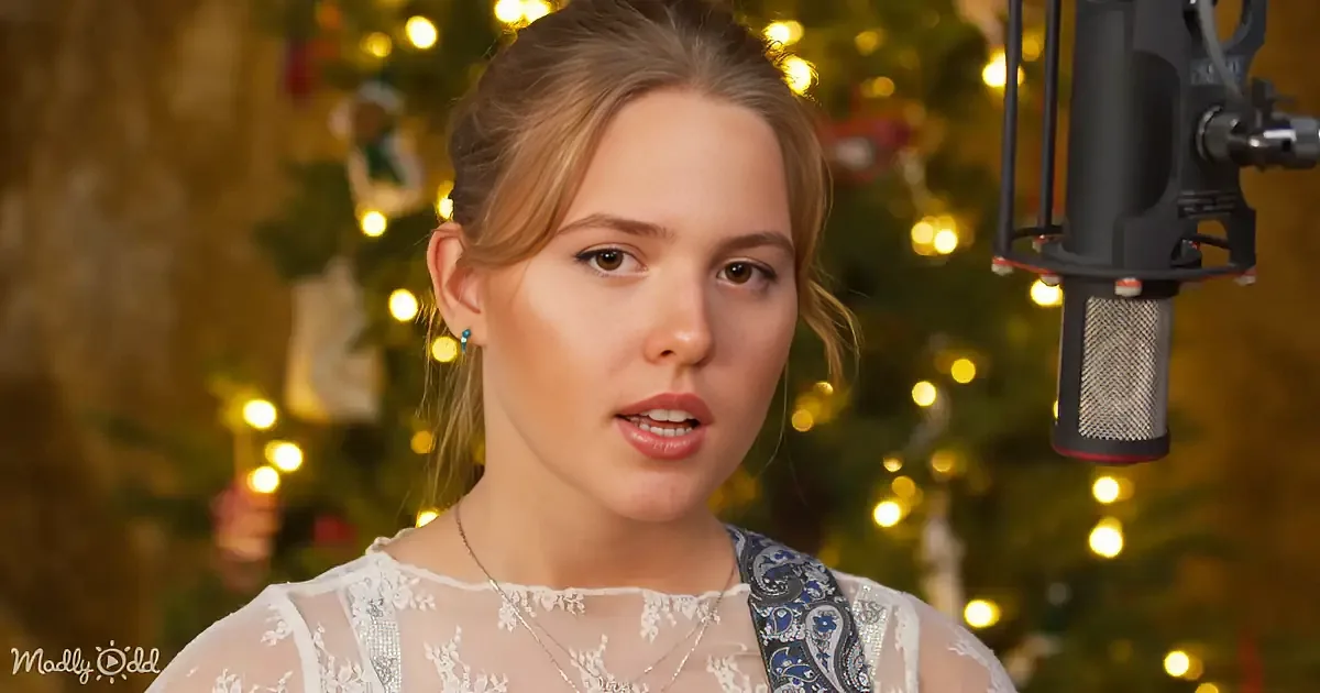 A woman in a white lace top speaks into a microphone, with a Christmas tree and lights in the background.