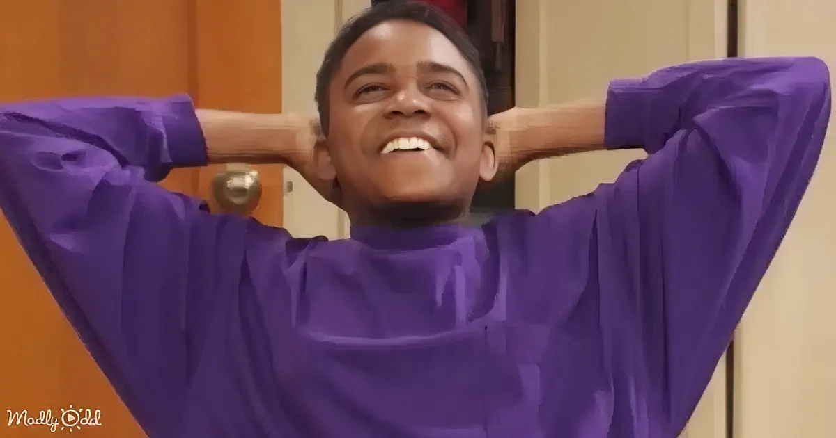 A person in a purple turtleneck shirt with hands behind their head, smiling.